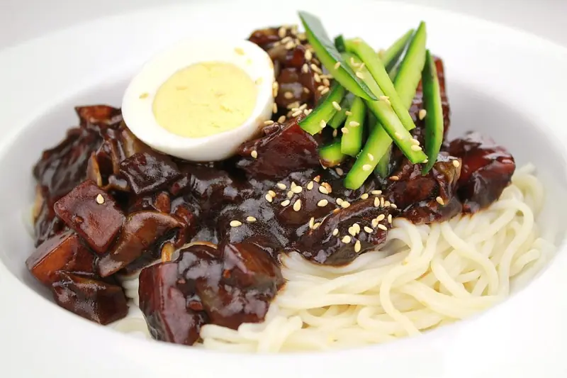 A plate of jjajangmyeon, Korean wheat noodles under glossy black bean sauce with pork, cucumber strips and a boiled egg