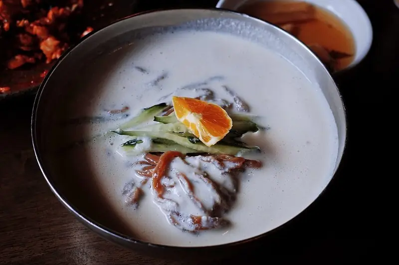 A bowl of kongguksu, chilled Korean wheat noodles in creamy white soy milk broth topped with cucumber and tomato