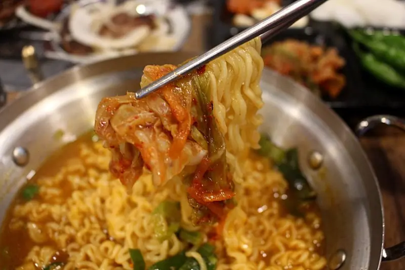 Korean instant ramyeon noodles lifted with chopsticks from a metal pot of spicy red broth, with napa cabbage kimchi alongside